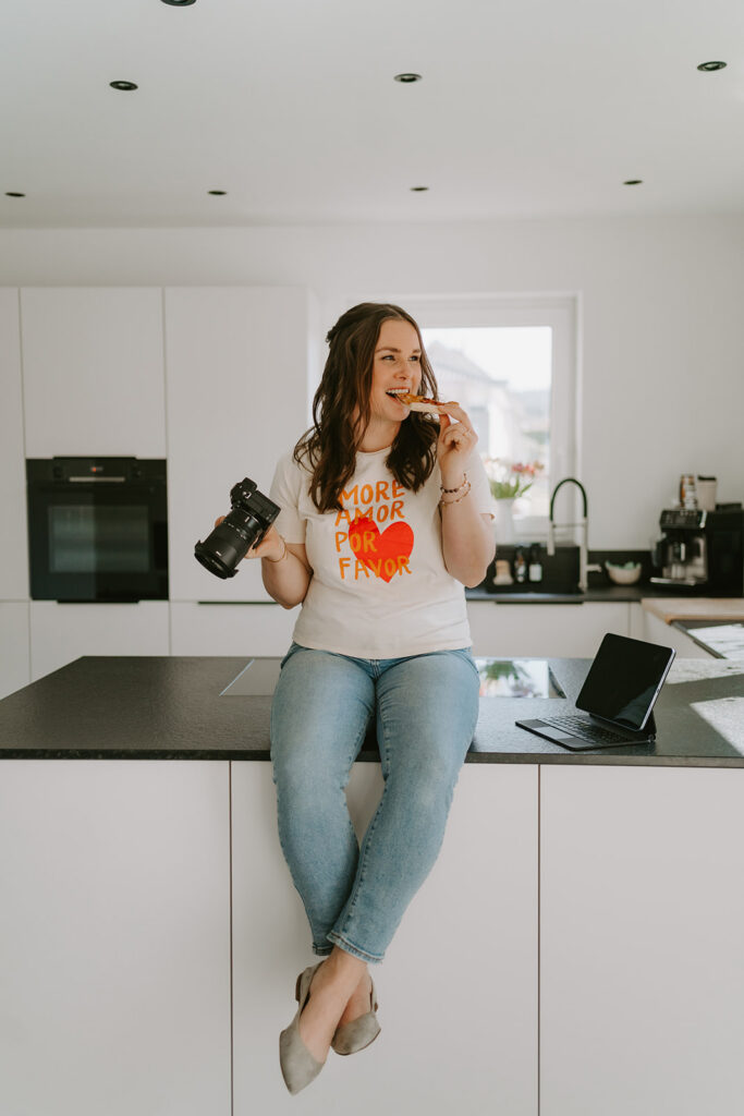 Food Fotografin Laura Fox sitzt auf der Küche mit Kamera in der Hand und beißt in ein Stück Pizza